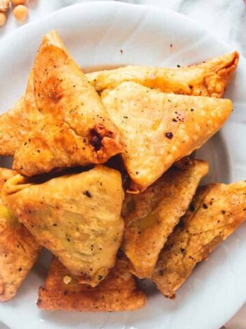 A white plate filled with golden-brown triangular pastries is placed on a marble surface. Around the plate are chickpeas, a sprig of cilantro, and some red peppers.