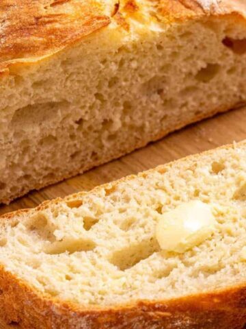A loaf of crusty bread with a slice cut off, resting on a wooden surface. The slice has a small pat of butter melting on top, highlighting the bread's airy crumb and golden crust.