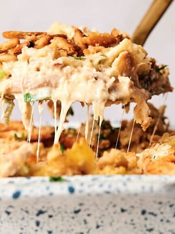 A close-up of a casserole being served from a speckled dish. The dish contains a mixture of creamy, cheesy ingredients topped with crispy golden breadcrumbs. A wooden spatula lifts a portion, revealing melted cheese strands.