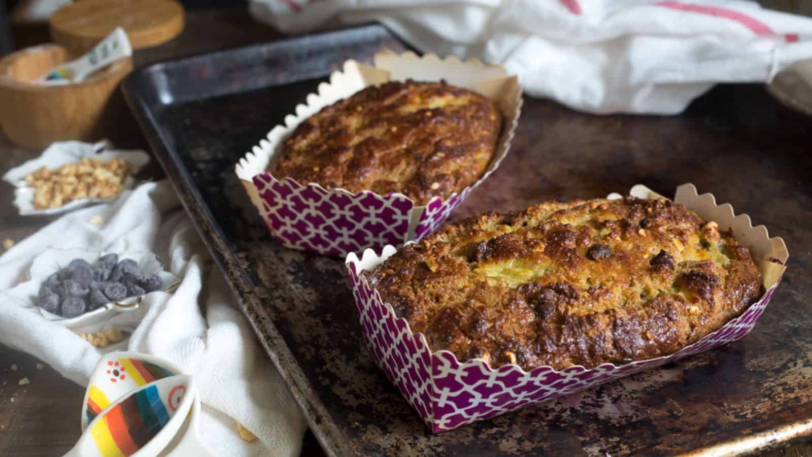 Two loaves of bread in decorative paper pans are on a baking tray. The bread is golden brown with a slightly textured surface. Nearby are small bowls containing chocolate chips and chopped nuts. A white cloth is partially visible in the background.