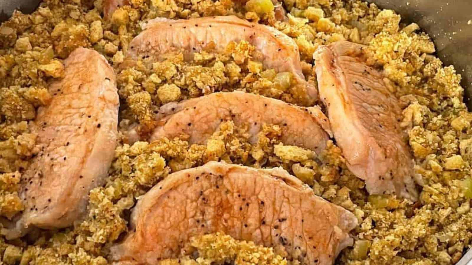 Five seasoned pork chops are arranged on a bed of golden-brown stuffing. The stuffing appears coarse with small pieces of celery visible. The dish is cooked and presented in a pan, showcasing a rustic and hearty meal.