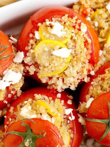 Five red tomatoes stuffed with quinoa, squash slices, and feta cheese are placed in a white baking dish. The tomatoes have been baked, with their tops cut off and positioned slightly to the side.