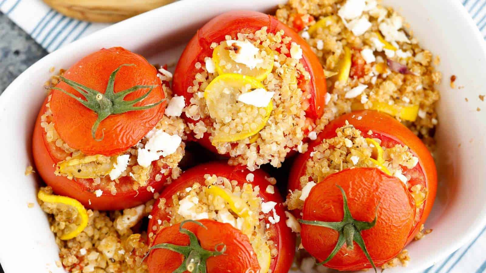 Five red tomatoes stuffed with quinoa, squash slices, and feta cheese are placed in a white baking dish. The tomatoes have been baked, with their tops cut off and positioned slightly to the side.
