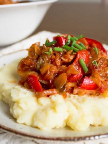 A plate of mashed potatoes topped with a red tomato-based sauce containing sliced bell peppers and garnished with chopped chives. The plate is on a patterned cloth, and there's a bowl in the background.
