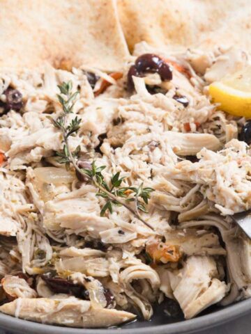 A plate of shredded chicken is garnished with herbs, black olives, and sliced cherry tomatoes. Two lemon wedges and pieces of flatbread accompany the dish. A fork is resting on the edge of the plate.