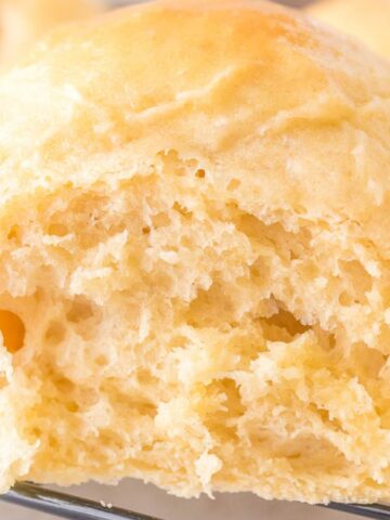 A close-up of a freshly baked bread roll on a cooling rack. The bread has a light golden crust and a soft, airy interior with visible texture. Additional rolls are partially visible in the background.