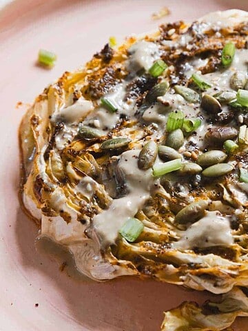 A roasted cabbage steak is on a light pink plate, topped with a drizzle of creamy sauce, seeds, and chopped green onions. The edges of the cabbage are charred, and the dish sits on a white surface.