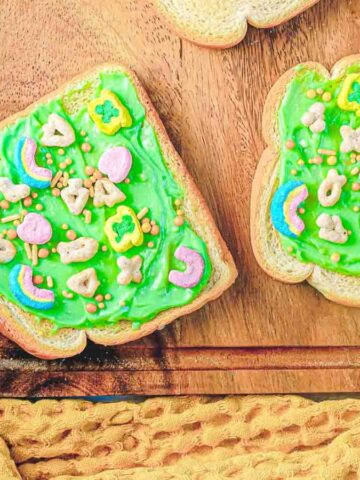 Two slices of bread with green icing and colorful cereal pieces on a wooden cutting board. A textured yellow cloth is partially visible on the table, alongside a bowl of cereal.