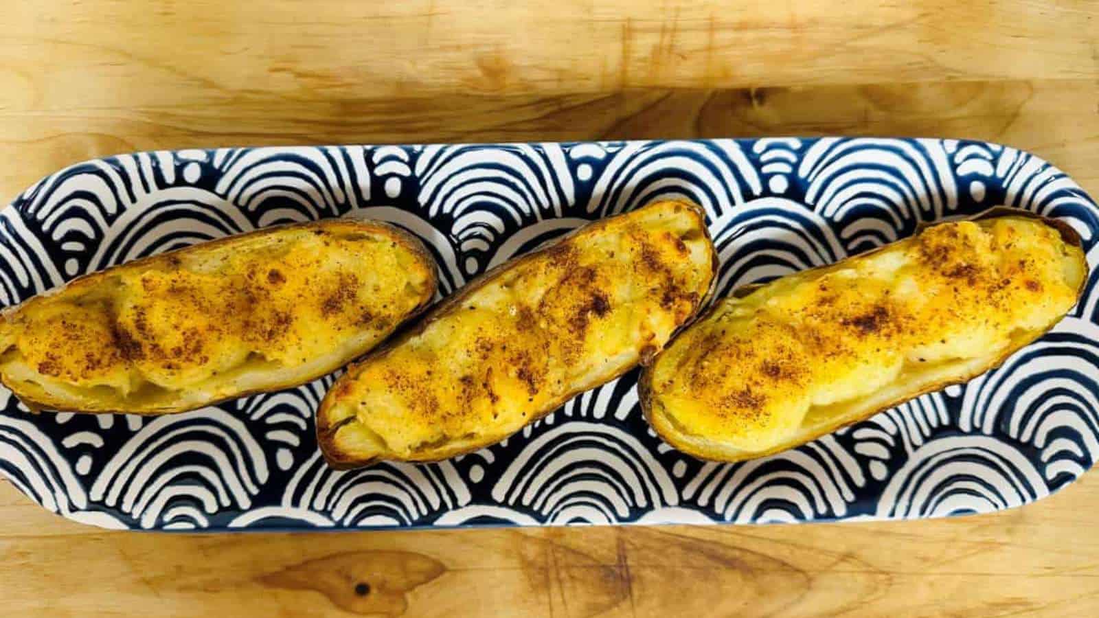 Three baked potato halves topped with melted cheese and a sprinkle of paprika are arranged in a blue and white patterned dish on a wooden surface.