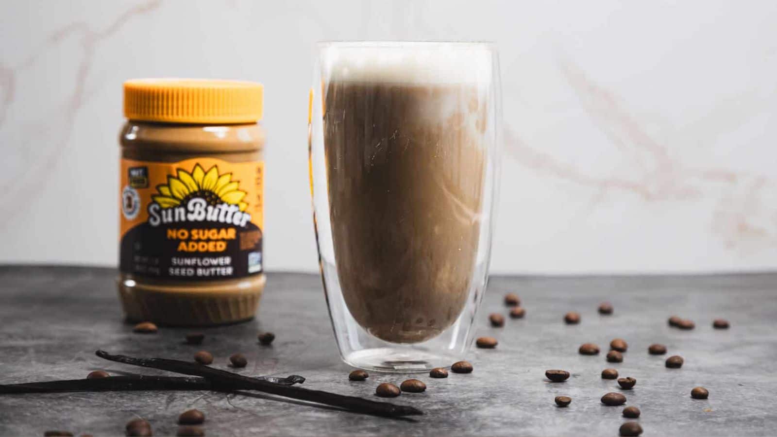 A glass of iced coffee with foam on top sits on a dark surface, surrounded by scattered coffee beans. A jar of SunButter 