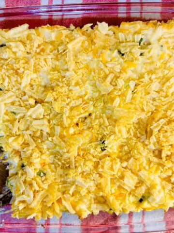 A classic comfort food casserole, baked to perfection in a glass dish and topped with crunchy crushed chips, with one hearty portion scooped out using a wooden spoon.