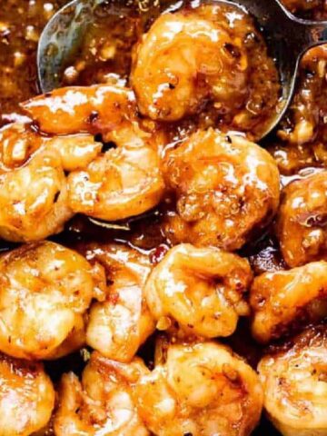 Sautéed shrimp in a glossy, brown garlic sauce are in a pan. A metal spoon is stirring the shrimp, which are coated in the sauce and positioned closely together. The dish appears to have spices and a rich texture.