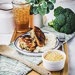 A plate with two browned chicken patties, reminiscent of a hearty stuffed chicken breast, sits atop mashed potatoes on a tray, accompanied by a bowl of shredded cheese. A glass of iced tea, a potted plant, and fresh broccoli are in the background, with a wooden spoon nearby.