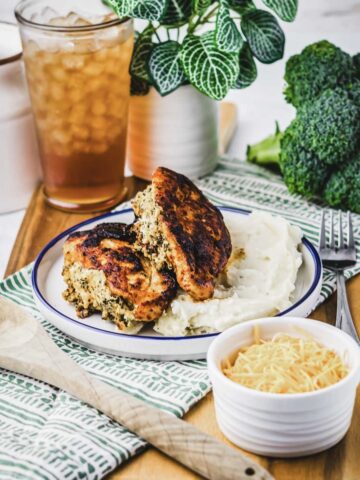 A plate with two browned chicken patties, reminiscent of a hearty stuffed chicken breast, sits atop mashed potatoes on a tray, accompanied by a bowl of shredded cheese. A glass of iced tea, a potted plant, and fresh broccoli are in the background, with a wooden spoon nearby.