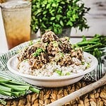 A white bowl filled with fluffy white rice is topped with Firecracker meatballs, drizzled with a creamy sauce and sprinkled with chopped green onions. It sits on a woven placemat, accompanied by a glass of iced drink and a plant in the background. Green onions garnish the scene.