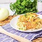 A plate with two servings of baked cheesy pasta topped with herbs sits on a white plate. The woven mat beneath holds a wooden spoon. In the background, an onion, parsley, and a purple cloth with white stripes evoke the comforting allure of a classic French Onion Chicken dish.
