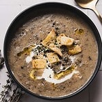 A bowl of creamy mushroom potato soup topped with croutons, a drizzle of olive oil, cracked pepper, and herbs. A small swirl of cream is visible. A gold spoon is placed next to the bowl on a white tiled surface, showcasing the delightful result of an Instant Pot soup masterpiece.