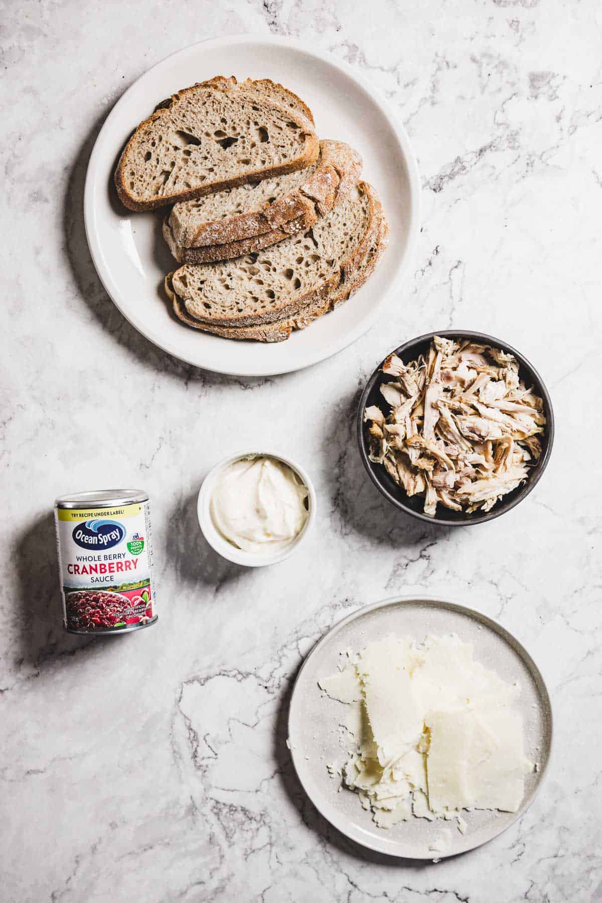 Slices of brown bread on a white plate, a bowl of shredded turkey, a can of whole berry cranberry sauce, a small bowl of white sauce, and a plate with cheese slices ready to melt, all set on a marble surface.