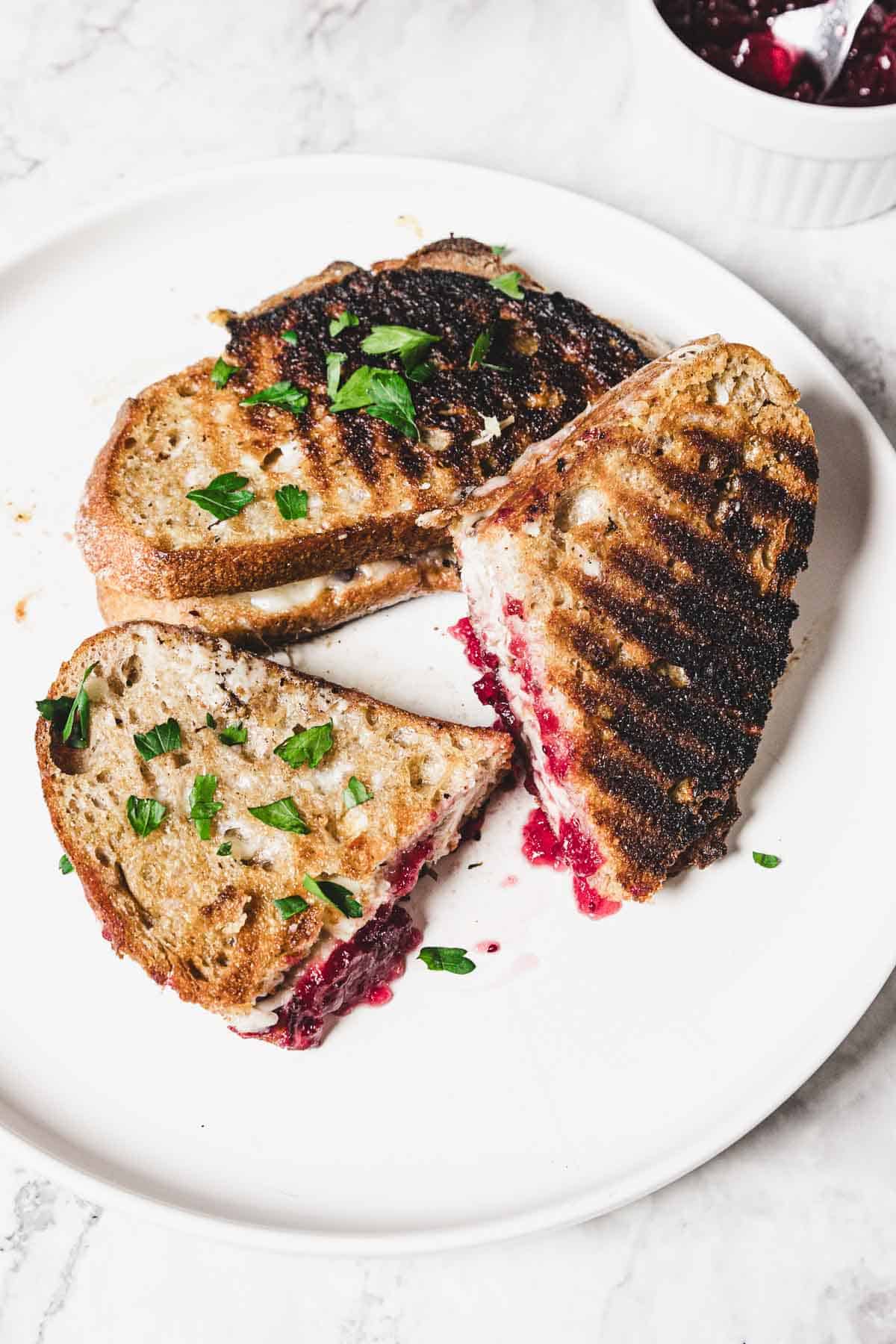 Three grilled sandwiches are on a white plate, each revealing a cross-section of melted cheese, turkey, and bright red cranberry jam. The sandwiches are garnished with small, fresh green herbs, and a bowl of extra cranberry jam awaits in the background on a marble surface.