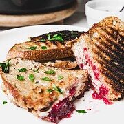 Grilled turkey melt halves rest invitingly on a white plate, bursting with cranberry sauce and other delicious ingredients. The dish is elegantly garnished with parsley, while a skillet and a small bowl of sauce linger in the softened background.