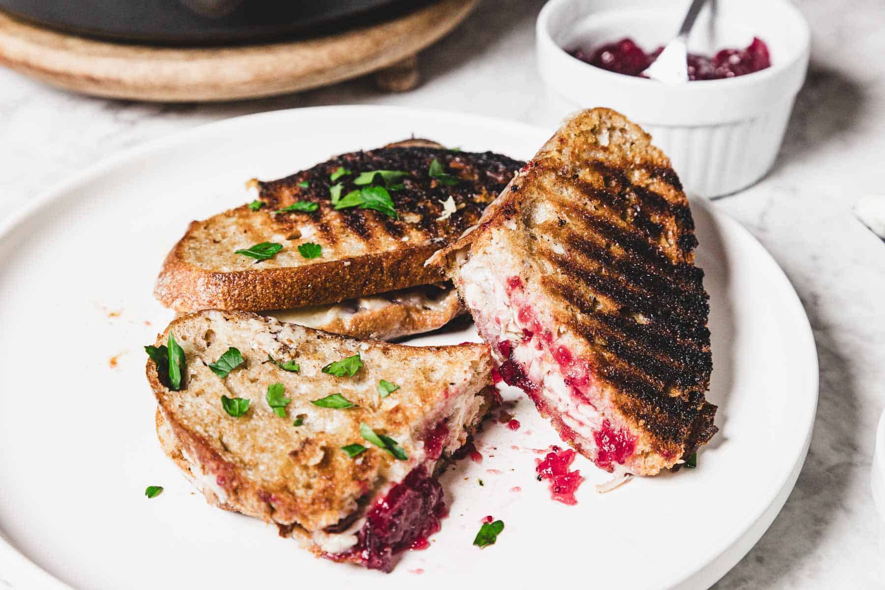 A turkey melt with grilled bread and cranberry filling is cut into quarters on a white plate. Green herbs garnish the sandwich. A small dish with red sauce and a spoon sits in the background on a marble surface.