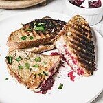 Grilled sandwiches on a white plate with melted turkey and cheese, filled with cranberry sauce. The bread sports grill marks and is sprinkled with chopped herbs. A small white dish containing extra cranberry sauce is in the background.