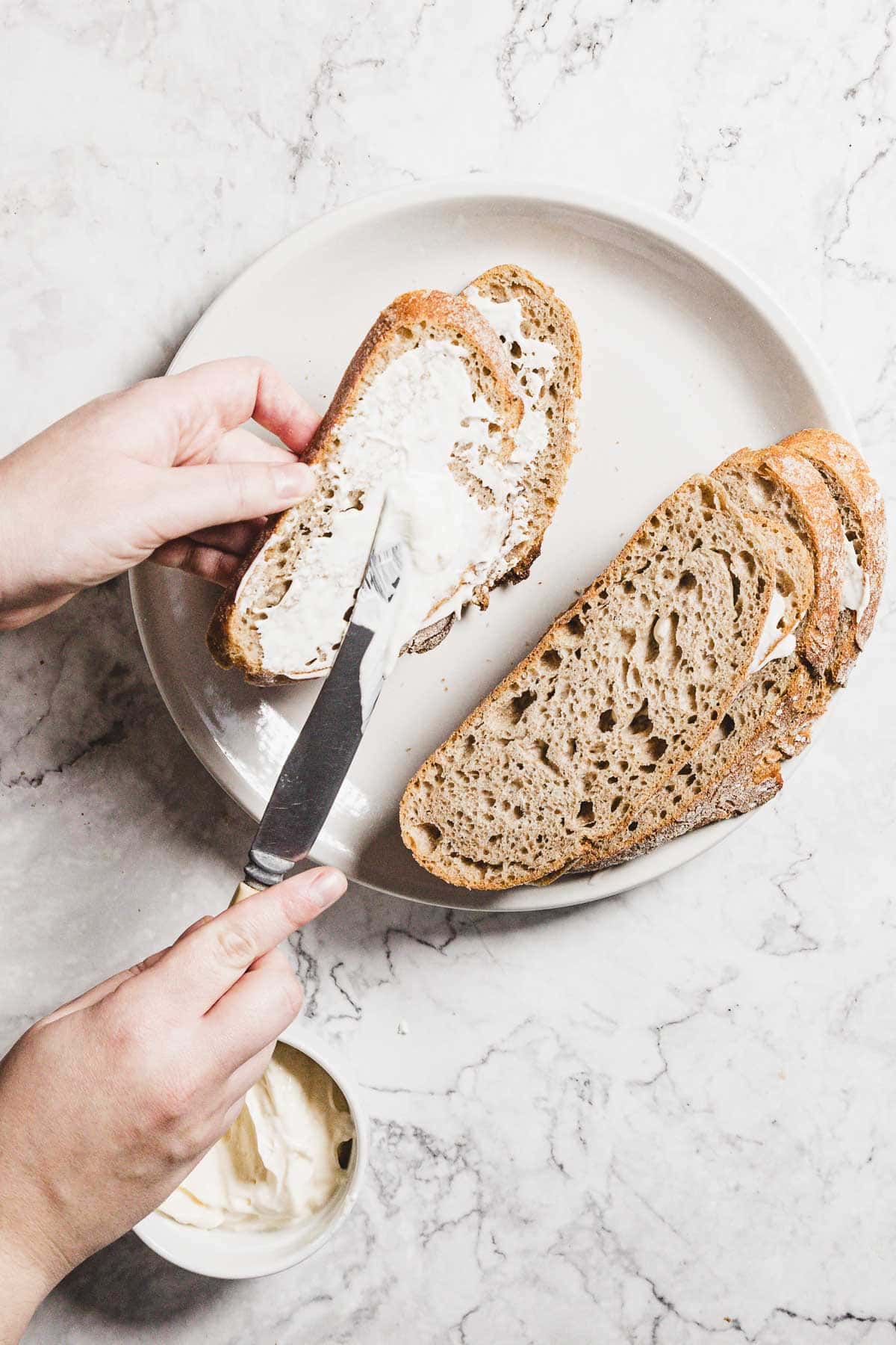 A person spreads butter on a slice of multigrain bread, preparing for a delightful turkey melt. The bread sits on a white plate atop a marbled surface. Nearby, a small bowl of butter awaits, with extra slices ready to transform into delicious cranberry-turkey creations.