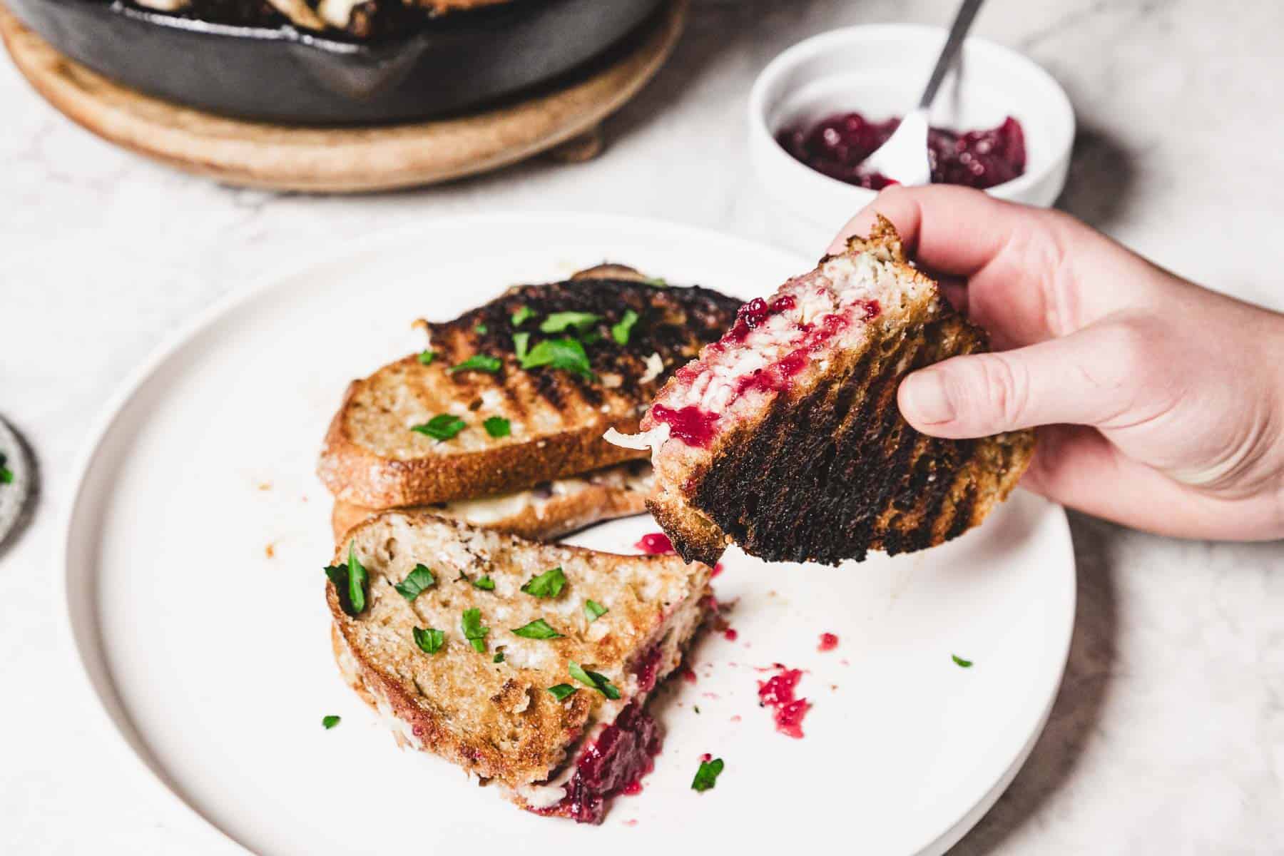 A grilled turkey melt with melted cheese and cranberry jam is being held over a white plate. The dish features two sandwich halves garnished with chopped parsley. In the background, there's a small bowl filled with extra cranberry jam.