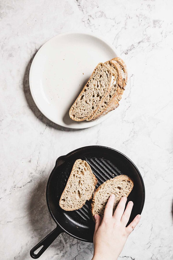 A hand carefully places a slice of bread into a black grill pan on the marble countertop, ready for a delicious melt. Nearby, a white plate holds several slices of cranberry-infused bread, waiting their turn for transformation.