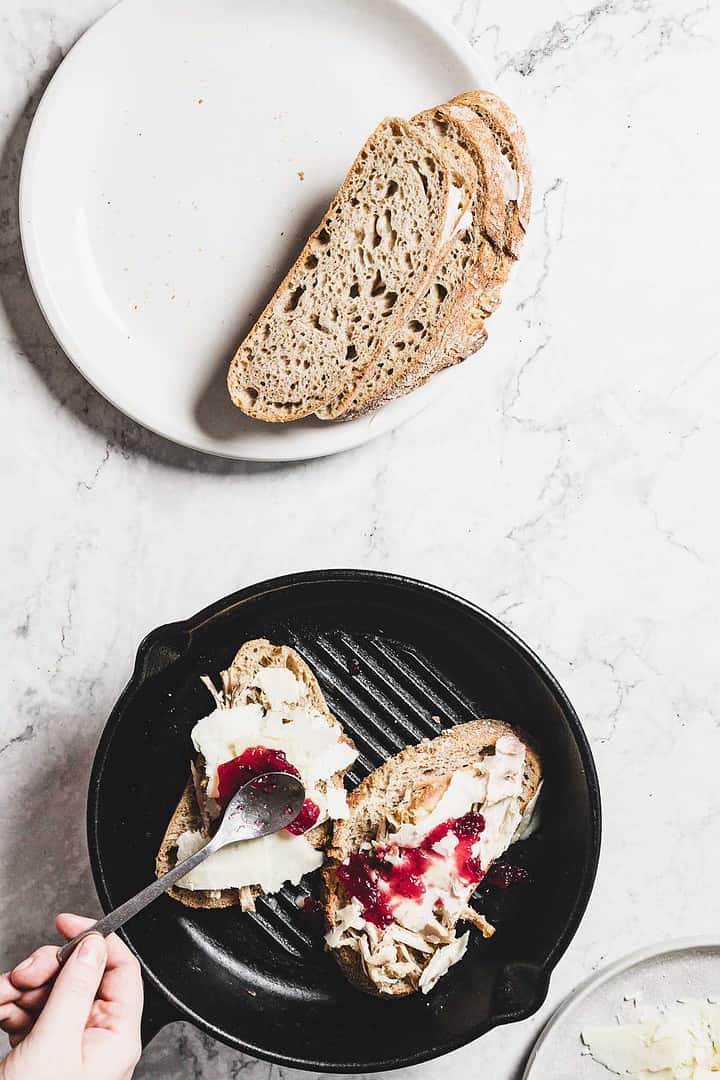 A hand spreads crimson cranberry jam on butter-topped bread slices in a black grill pan. Above, a white plate cradles two more slices. The marble surface serves as the perfect backdrop for this delightful scene.