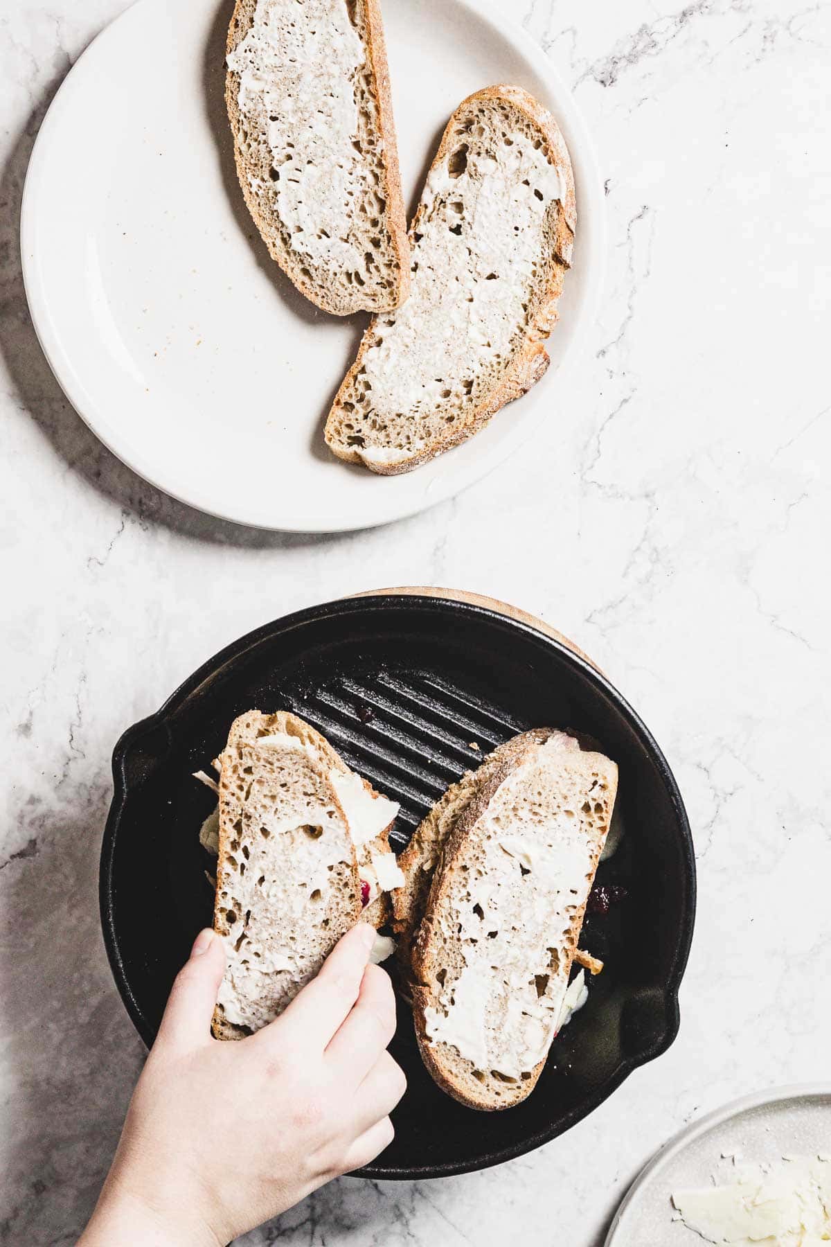 A hand places a slice of buttered bread onto a black grill pan, ready for the melt. Two other slices with cranberry and butter are on a white plate nearby. The background is a light marble surface.