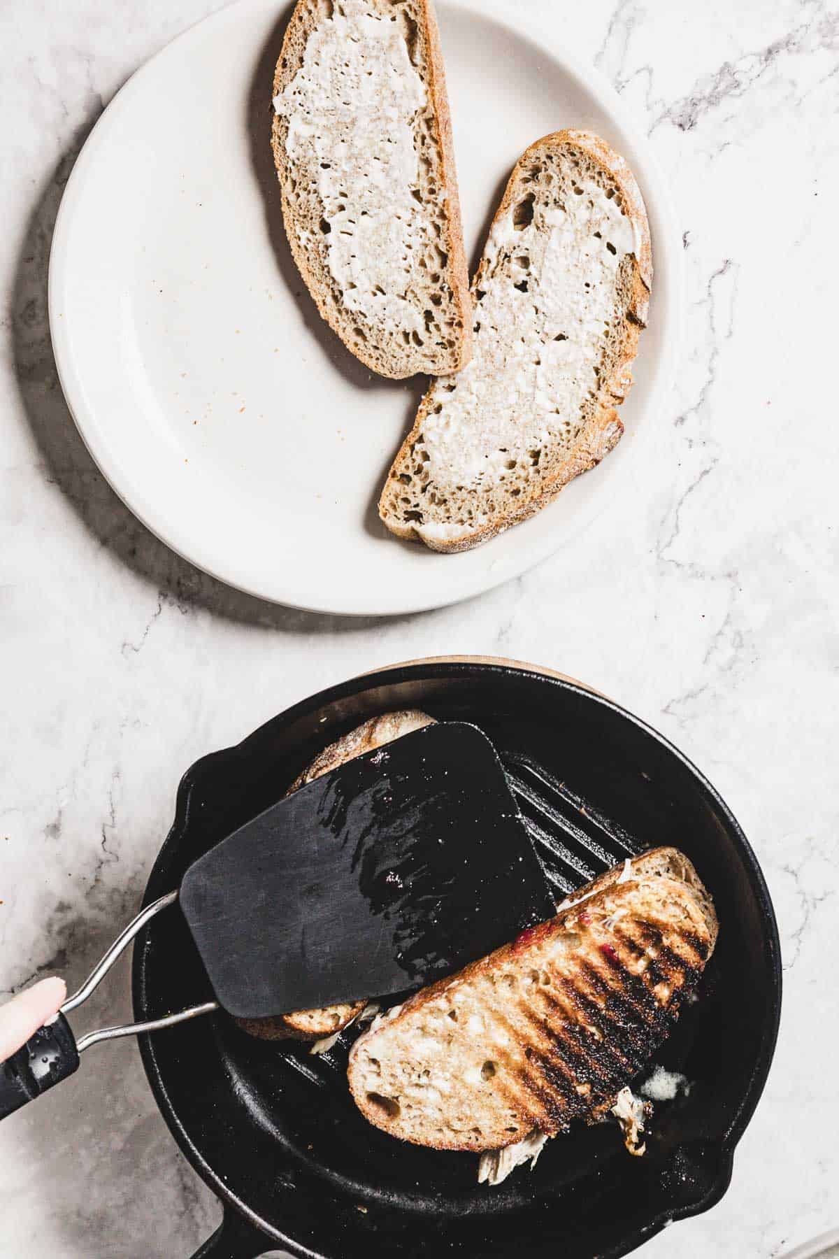 A person holds a spatula, pressing down on a turkey melt in a cast iron skillet. Two slices of bread with butter rest on a white plate in the background, set on the white marble surface. A hint of cranberry peeks from the sandwich's edge, promising a sweet tang with each bite.