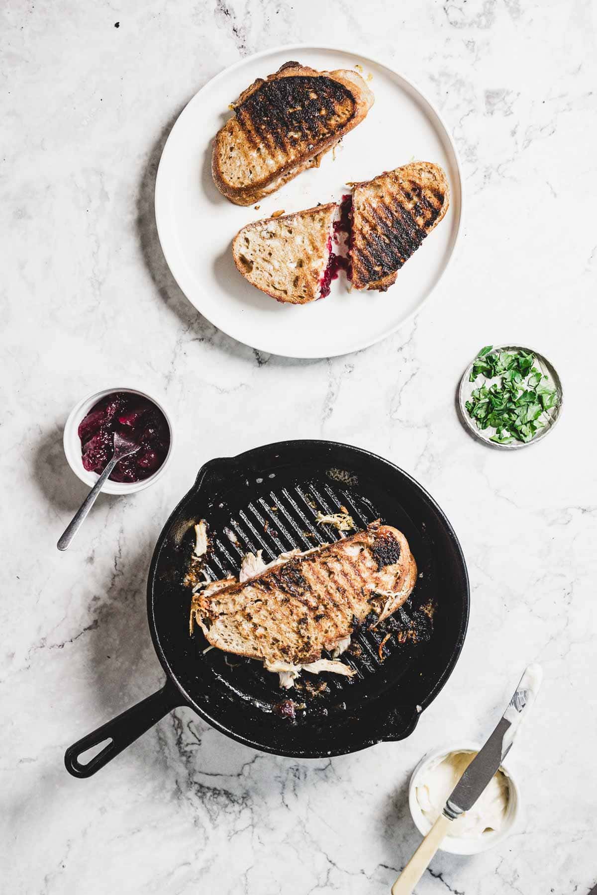 A grilled cheese sandwich melts in a skillet, with gooey cheese oozing out. The skillet rests on white marble. Nearby, a sliced sandwich with turkey sits on a plate beside a small bowl of cranberry jam and chopped greens.