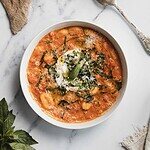 A bowl of tomato-based gnocchi alla vodka topped with grated cheese and shredded basil sits on a marble surface. A dollop of cream is in the center, and a spoon rests next to the bowl. Fresh basil leaves are nearby.