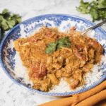 A blue and white bowl with a serving of shredded chicken curry tops fluffy white rice, garnished with vibrant cilantro leaves. A silver spoon rests in the bowl, hinting at a cozy slow cooker recipe. Fresh cilantro is scattered on the marble surface beside the bowl, near a mustard-colored cloth.