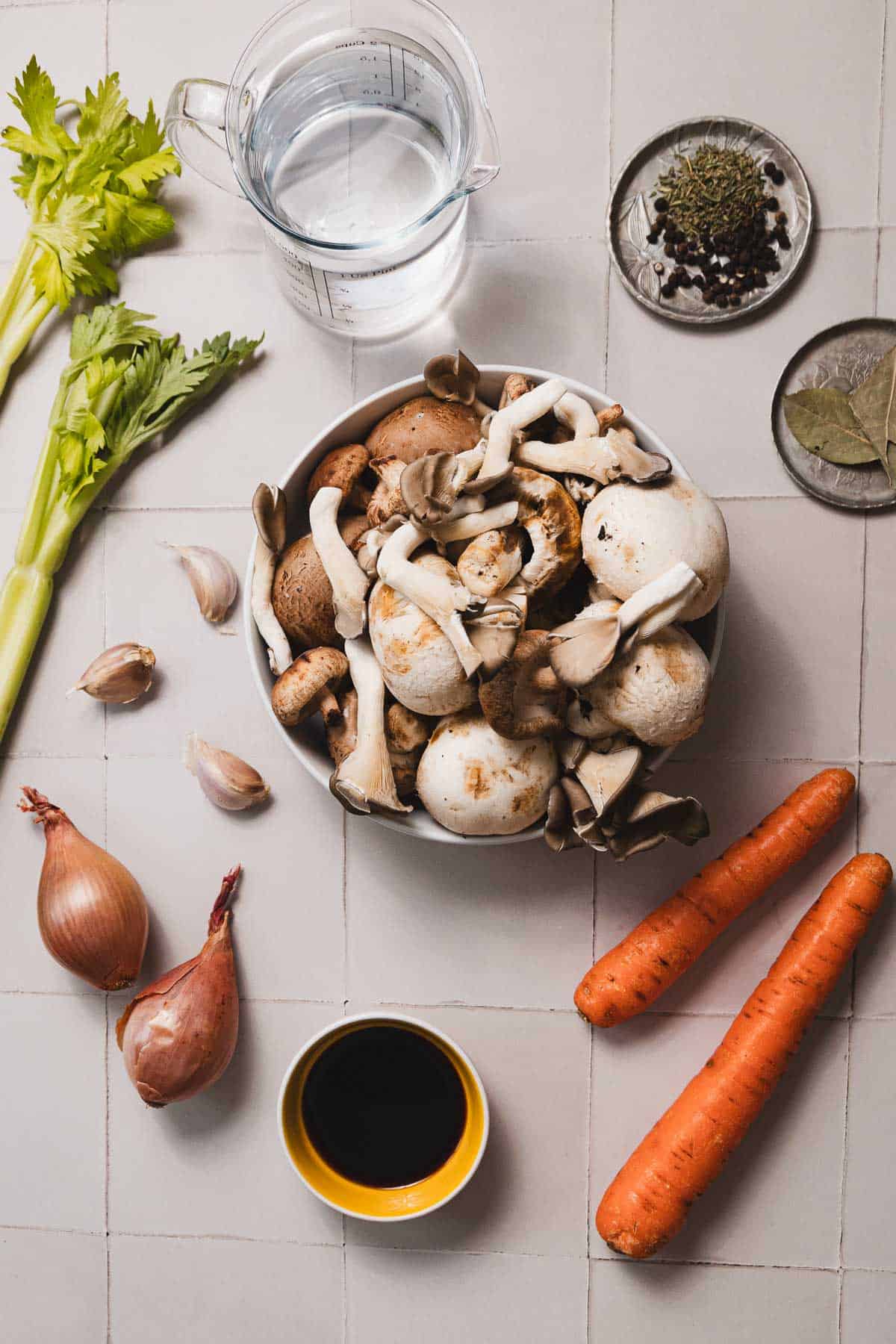 A tiled surface displays celery stalks, a measuring cup of water, and ingredients for a savory mushroom broth: a bowl of various mushrooms, spices on a small plate, bay leaves, garlic cloves, shallots, soy sauce in a small bowl, and two carrots. Perfect for your next slow cooker recipe.