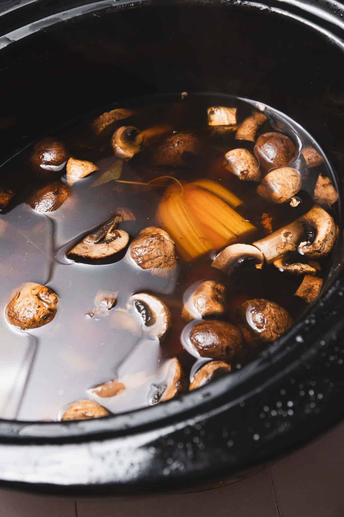 A black slow cooker sits on the countertop, brimming with a rich mushroom broth recipe. Within the dark liquid, brown mushrooms and chunks of orange spaghetti squash float gently, creating an aromatic blend that's sure to satisfy any comfort food craving.