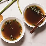 Two white bowls filled with clear mushroom broth, perfected in a slow cooker, are garnished with sliced green onions. The bowls rest on a light-colored tiled surface, with a wooden spoon nearby and fresh green stalks beside them.