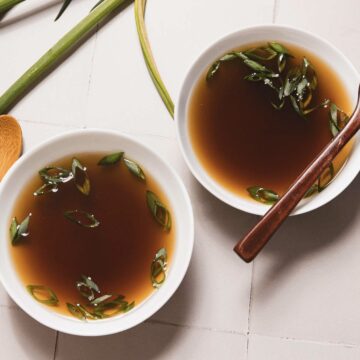 Two white bowls filled with clear mushroom broth, perfected in a slow cooker, are garnished with sliced green onions. The bowls rest on a light-colored tiled surface, with a wooden spoon nearby and fresh green stalks beside them.