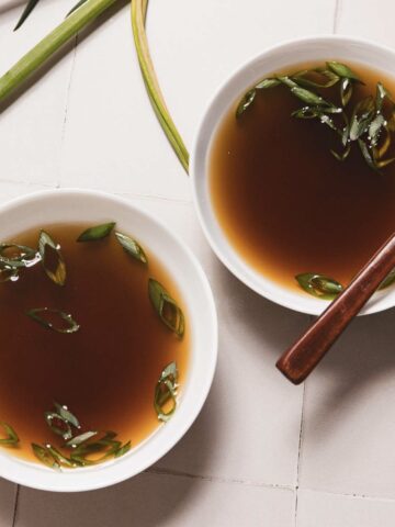 Two white bowls filled with clear mushroom broth, perfected in a slow cooker, are garnished with sliced green onions. The bowls rest on a light-colored tiled surface, with a wooden spoon nearby and fresh green stalks beside them.