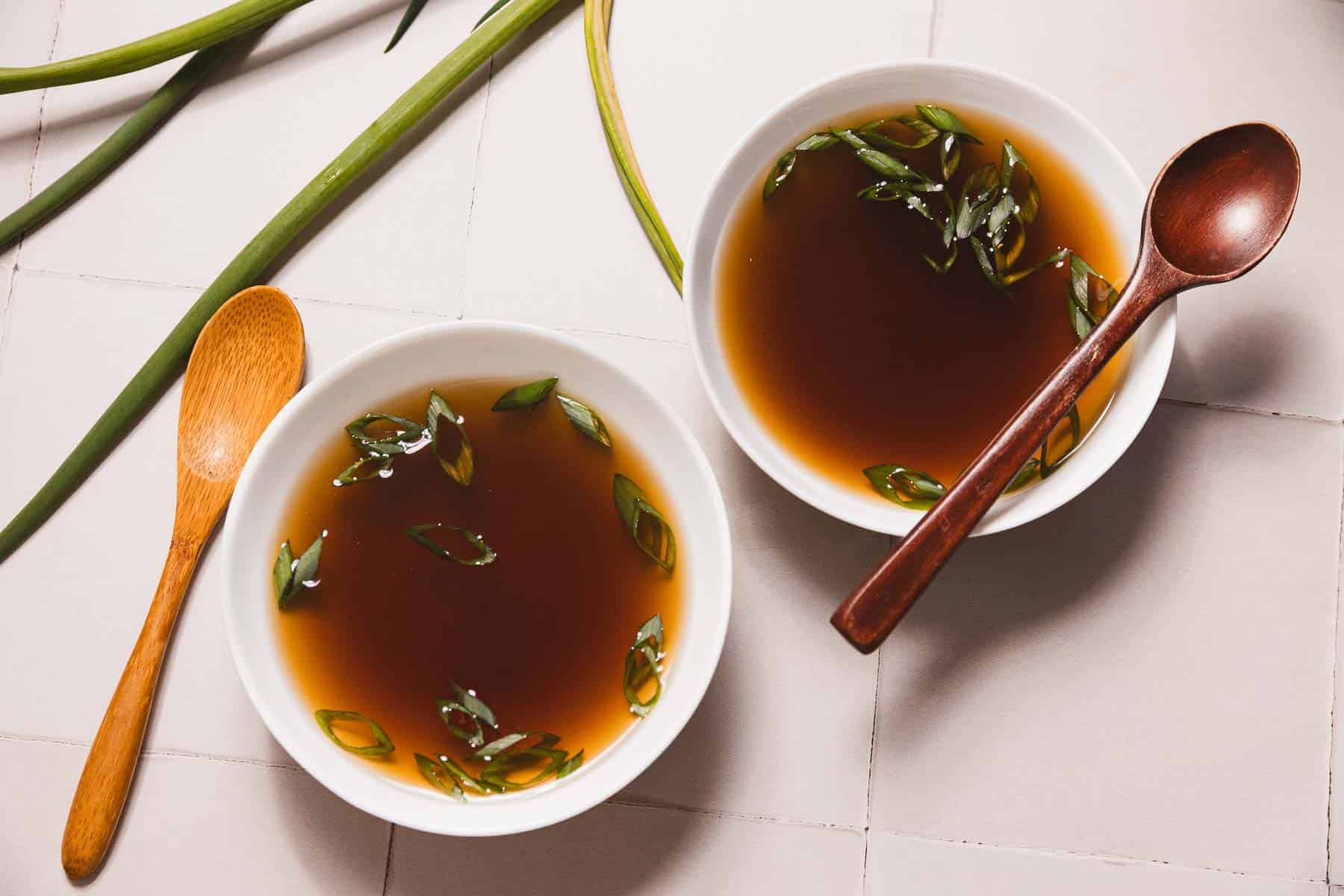 Two white bowls filled with a rich mushroom broth are garnished with sliced green onions, resting on a light surface. A wooden spoon is positioned beside each bowl. Green onion stalks peek from the top left corner, inviting you to savor this slow-cooker recipe.