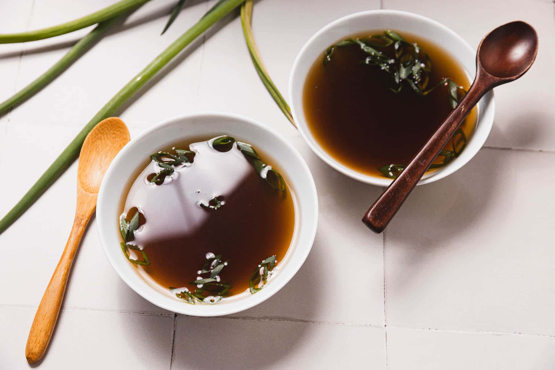 Two bowls of miso soup with green onion slices rest on a white tiled surface, their flavors enhanced by a rich mushroom broth. Each bowl is paired with a wooden spoon, and green onion stalks are artfully arranged in the background.