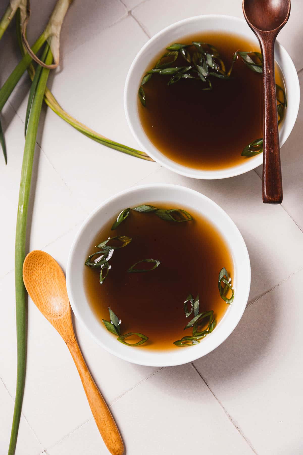 Two white bowls of aromatic mushroom broth, garnished with fresh green onions, rest on a tiled surface. This easy recipe gives a homely touch when made in a slow cooker. Wooden spoons lie beside the bowls, and vibrant green onion stalks peek from the corner.