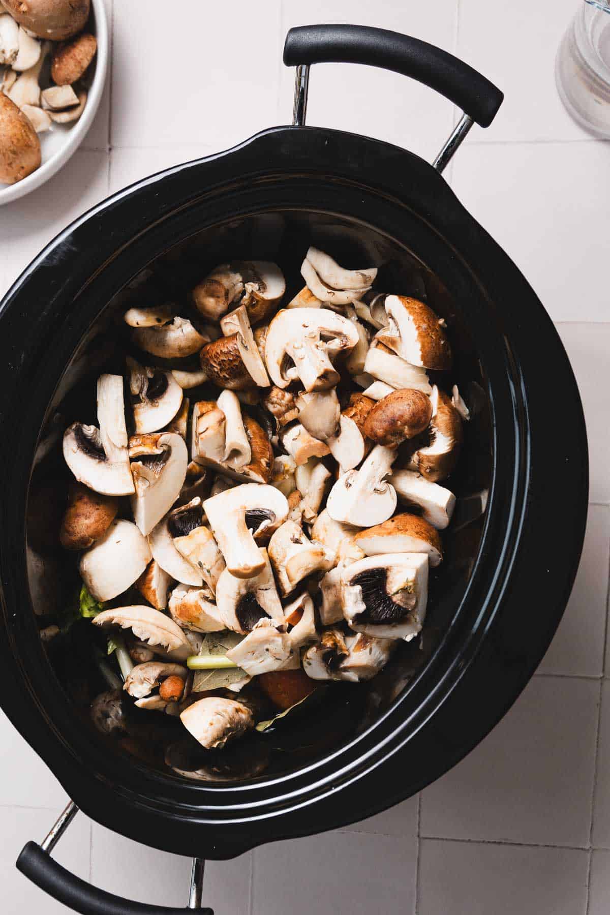 A slow cooker filled with sliced mushrooms is nestled on a white tiled surface, preparing a savory mushroom broth. A bowl of additional mushrooms peeks in from the top left corner. The slow cooker, featuring a black lid with metal handles, promises a delicious recipe in the making.