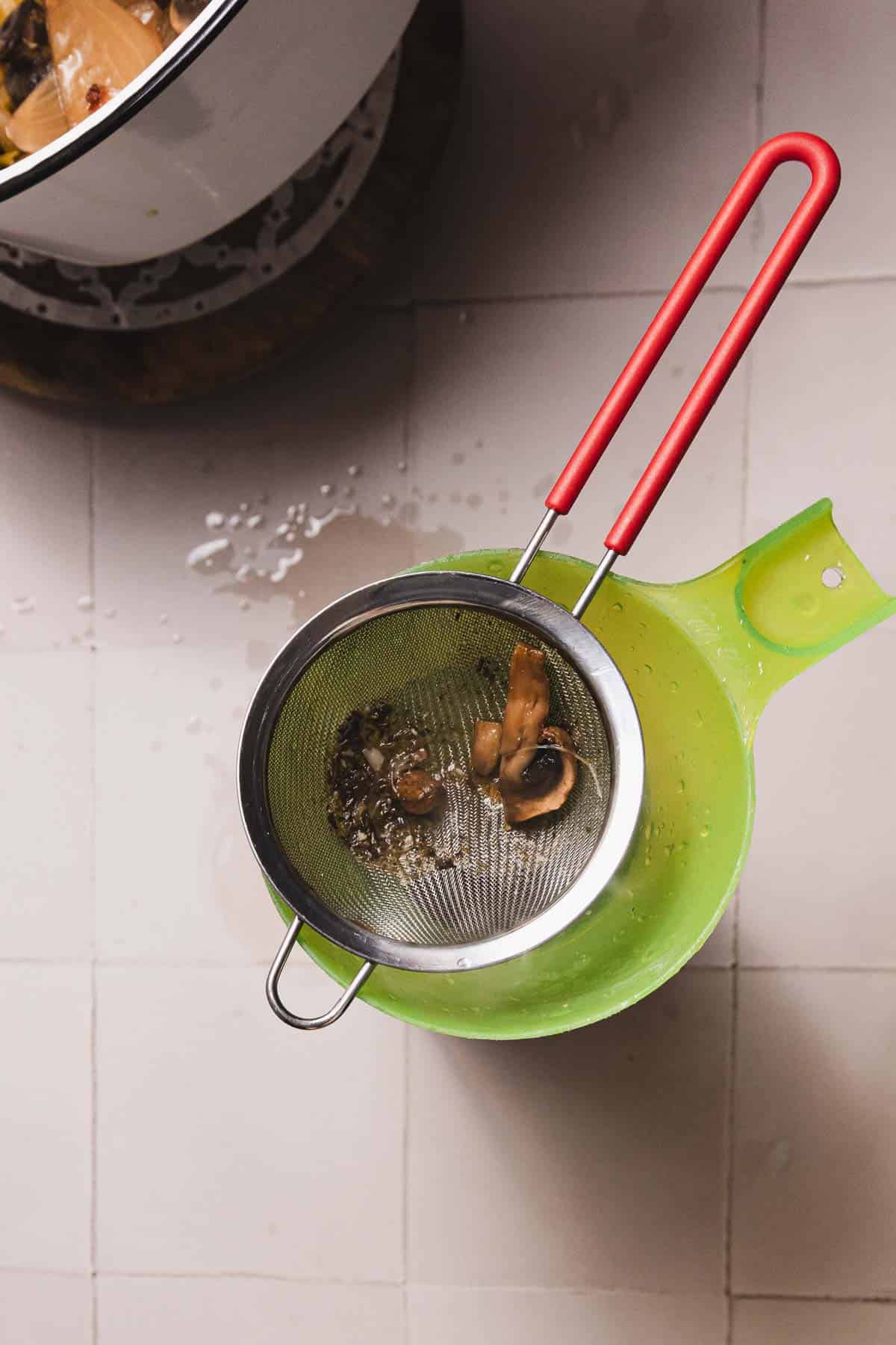 A metal strainer with a red handle rests over a light green bowl on a tiled surface. Inside, small mushroom pieces hint at an upcoming recipe. A cookware pot peeks from the top left, while water droplets speckle the tiles, evoking thoughts of a simmering mushroom broth.
