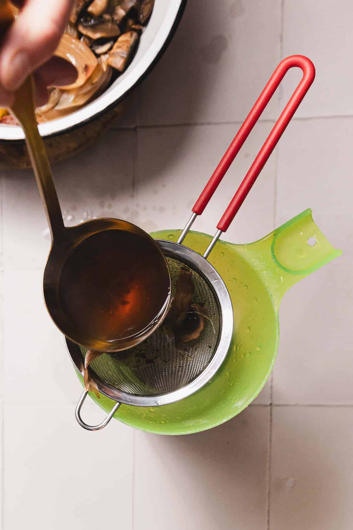A person pours mushroom broth from a ladle through a metal strainer into a green container. The strainer rests securely, with mushroom pieces visible. In the background on the tiled surface, a bowl holds leftover scraps, hinting at an intricate recipe in progress.