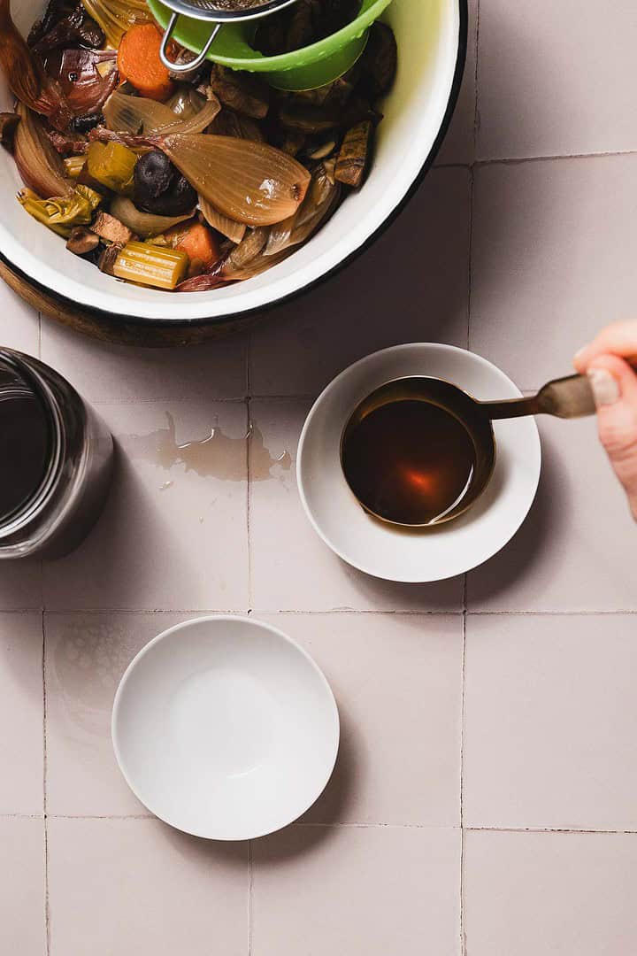 A person pours soothing mushroom broth from a ladle into a small white bowl. Nearby, a slow cooker brims with various cooked vegetables, and another empty bowl rests on the tiled surface. A glass jar completes this cozy culinary scene.