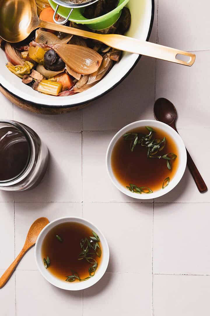 Two bowls of mushroom broth garnished with green onions rest on a light tile surface. Beside them is a jar, a spoon, and a plate filled with fresh vegetables and a wooden spatula&mdash;perfect for those slow cooker days when you savor every moment of the recipe.