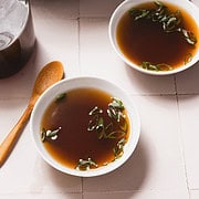 Two bowls of mushroom broth, garnished with sliced green onions, rest on a tiled surface. A wooden spoon lies nearby, and a glass jar of broth is partially visible. In the background, a casserole dish with vegetables hints at the delicious recipe made in a slow cooker.