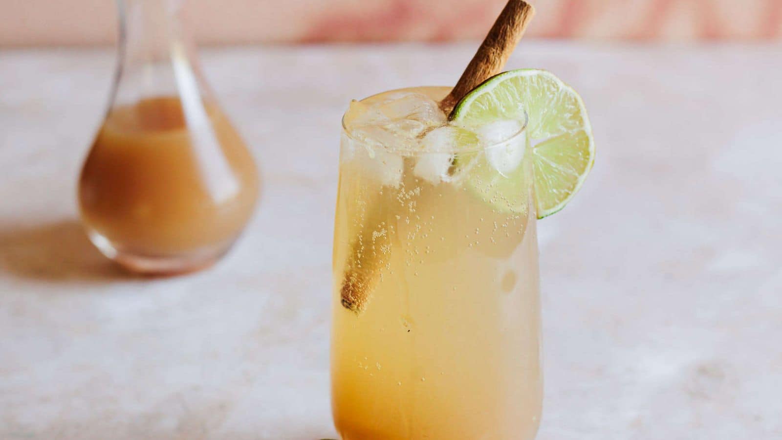 A glass filled with a light brown iced drink sits on a surface. The drink is garnished with a lime slice and a cinnamon stick. In the background, there&rsquo;s fresh ginger, half a lime, and a bottle partially visible.
